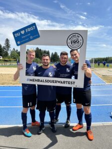 Vier Fußballspieler posieren zusammen mit einem großen Rahmen auf einem Sportplatz.