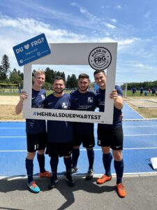 Vier Fußballspieler posieren zusammen mit einem großen Rahmen, der den Text 'DU FRG?' enthält.