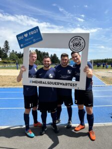 Vier Fußballspieler posieren zusammen mit einem großen Rahmen, der den Text 'DU ♥ FRG?' enthält.