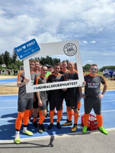 Eine Fußballmannschaft posiert mit einem Rahmen und einem Plakat auf einem Sportplatz.