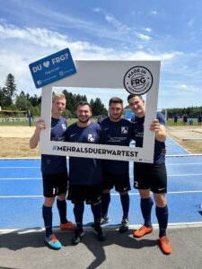 Vier Fußballspieler posieren zusammen mit einem großen Rahmen auf einem Sportplatz.