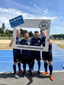 Vier Spieler einer Fußballmannschaft posieren mit einem großen Rahmen auf dem Spielfeld.