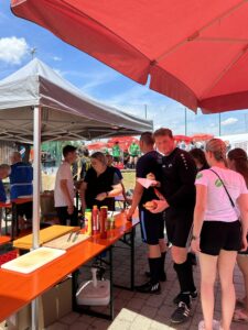 Personen stehen an einem Imbissstand mit Tischen und Sonnenschirmen während eines Sportevents.