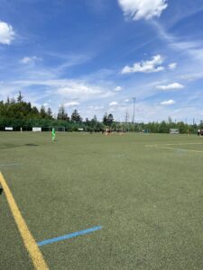 Fußballfeld mit Spielern und Torwart auf einem Kunstrasenplatz unter blauem Himmel.