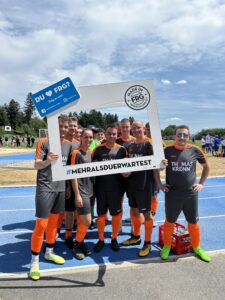Gruppe von Fußballspielern in grauen Trikots mit orangefarbenen Akzenten, die einen Rahmen halten.