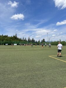 Gruppenspiel auf einem Kunstrasenplatz mit Spielern in verschiedenen Trikots und einer Zuschauergruppe im Hintergrund.