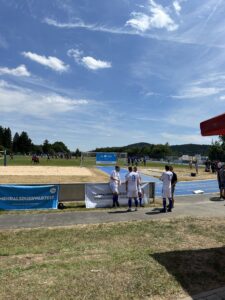 Drei Fußballspieler in weißen Trikots stehen auf einem Sportplatz mit Sandfeld im Hintergrund.