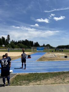 Zwei Fußballspieler in blauen Trikots stehen auf einem Sportplatz mit einem Fußballtor im Hintergrund.