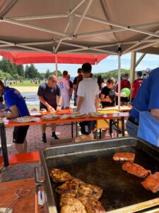 Grillstation mit gegrilltem Fleisch und Personen, die Essen zubereiten und servieren.