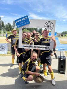 Gruppe von Fußballspielern posiert mit einem großen Rahmen und einem Schild bei einem Sportereignis.