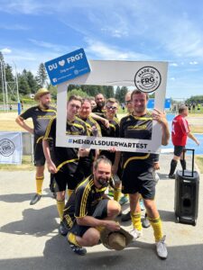 Gruppe von Fußballspielern posiert mit einem großen Rahmen und einem Schild auf einem Sportplatz.