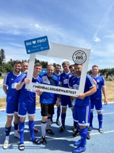 Eine Gruppe von Fußballspielern in blauen Trikots posiert mit einem großen Rahmen auf einem Sportplatz.