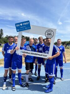Eine Gruppe von Fußballspielern in blauen Trikots posiert mit einem großen Rahmen auf einem Sportplatz.