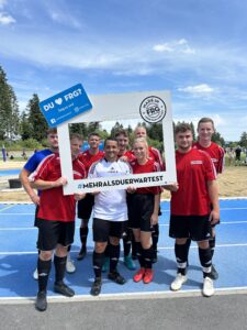 Eine Gruppe von sieben Fußballspielern in roten Trikots posiert mit einem großen Rahmen auf einem Sportplatz.