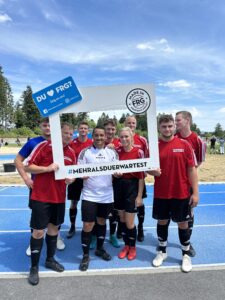 Eine Gruppe von Fußballspielern in roten Trikots posiert mit einem großen Rahmen auf einem Sportplatz.