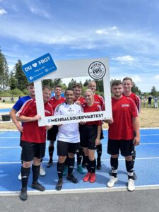 Eine Gruppe von Fußballspielern in roten Trikots posiert mit einem großen Rahmen und einem Hashtag.