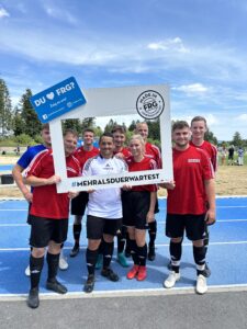 Eine Gruppe von sieben Personen in roten Fußballtrikots posiert mit einem Rahmen auf einem Sportplatz.