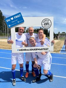 Vier Männer in Fußballtrikots posieren mit einem großen Rahmen auf einem Sportplatz.