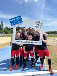 Eine Gruppe von acht Männern in Fußballtrikots posiert mit einem Schild, das den Hashtag #MEHRALSDUERWARTEST zeigt.