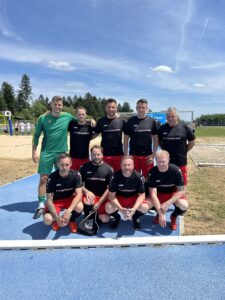 Eine Fußballmannschaft in schwarzen und roten Trikots steht auf einem Sportplatz mit Toren im Hintergrund.