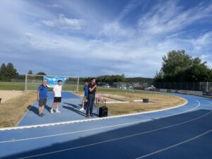 Drei Personen stehen auf einer Laufbahn bei einer Sportveranstaltung, während eine Person spricht.