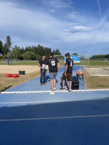 Drei Personen in Sportkleidung stehen auf einem Sportplatz mit einem Fußballtor im Hintergrund.