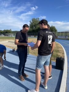 Zwei Männer stehen auf einem Sportplatz und diskutieren über Dokumente in der Hand.
