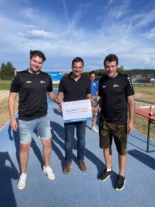 Drei Männer stehen auf einem Sportplatz und halten ein Schild mit der Aufschrift '30 Liter Freibier'.