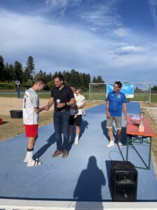 Ein Fußballspieler in Shorts und T-Shirt erhält eine Auszeichnung von einem Mann, während ein Kind gehalten wird.