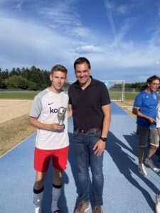 Junger Fußballspieler in Sportkleidung hält einen Pokal zusammen mit einem Erwachsenen auf einem Sportplatz.