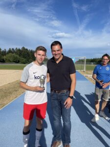 Junger Fußballspieler in einem weißen Trikot mit Pokal, stehend neben einem Mann in einem schwarzen T-Shirt.