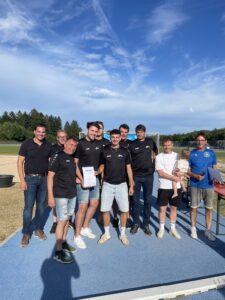 Gruppe von Männern in schwarzen T-Shirts, die einen Pokal halten, umgeben von weiteren Personen im Freien.