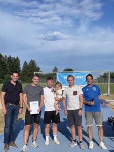 Fünf Personen stehen auf einem Sportplatz, eine Person hält ein Kind und eine Urkunde.