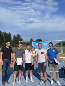 Fünf Personen stehen auf einem Sportplatz, zwei halten Urkunden, eine Person hält ein Kind.