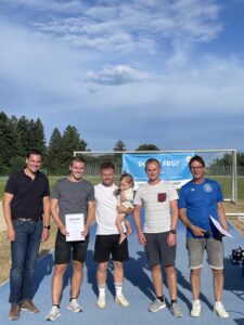 Fünf Personen stehen auf einem Sportplatz und halten Urkunden in den Händen, während ein Kind auf einem der Männer sitzt.