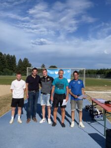 Fünf Personen stehen auf einem Sportplatz vor einem Fußballtor und halten Dokumente in der Hand.