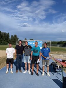 Fünf Personen stehen auf einem Sportplatz, im Hintergrund ein Fußballtor und ein Banner.