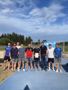 Eine Gruppe von acht Männern steht auf einem Sportplatz, einige tragen Fußballtrikots und Shorts.