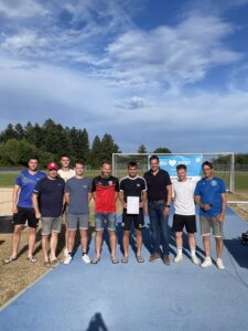 Gruppe von neun Fußballspielern, die eine Urkunde in der Mitte halten, auf einem Sportplatz.