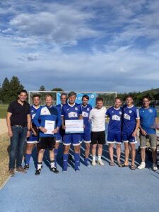 Eine Fußballmannschaft in blauen Trikots steht zusammen mit Urkunden auf einem Sportplatz.