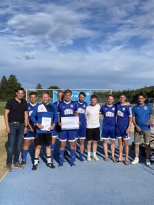 Eine Fußballmannschaft in blauen Trikots hält eine Urkunde auf einem Sportplatz.