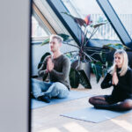 Zwei Personen sitzen im Schneidersitz auf Yogamatten und meditieren in einem Raum mit großen Fenstern.