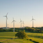 Sechs Windkraftanlagen stehen in einer grünen Landschaft unter blauem Himmel.