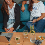 Zwei Frauen sitzen auf einer Couch mit Getränken und Laptops auf einem Tisch.