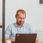 Ein Mann mit Bart sitzt an einem Tisch und arbeitet an einem Laptop. Eine Tasse steht neben ihm.