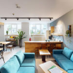 Moderne Wohnküche mit blauen Sofas, Holztisch und Küchenzeile mit Pflanzen im Hintergrund.