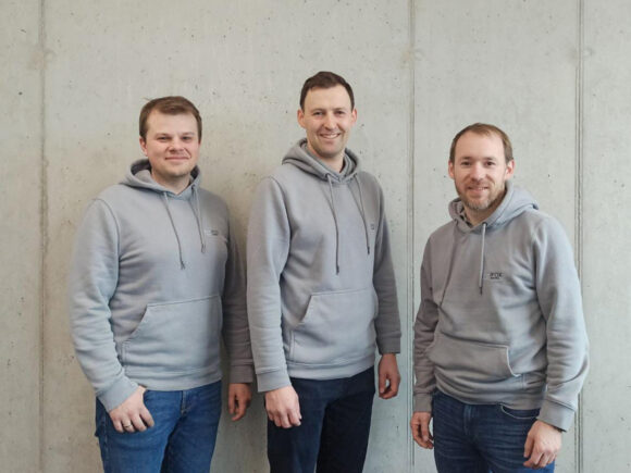 Drei Männer stehen nebeneinander in grauen Hoodies vor einer Betonwand.