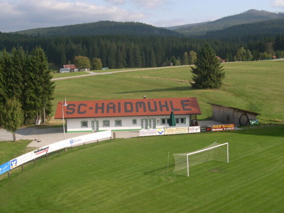 Vereinsheim des SC Haidmühle mit großem Schriftzug auf dem Dach und Fußballfeld im Vordergrund.