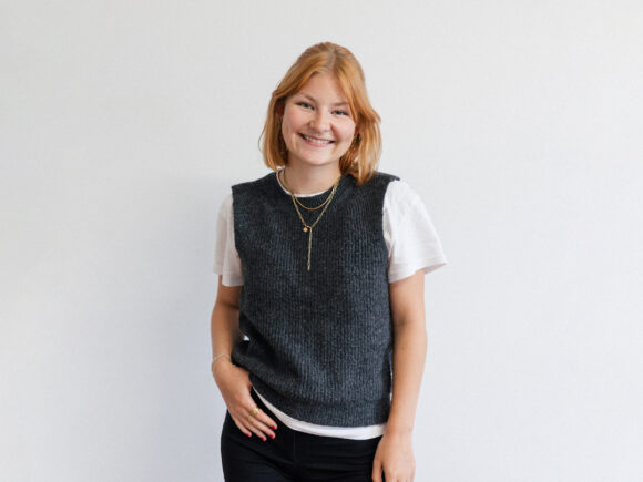 Eine Frau mit blonden Haaren und einem grauen Pullover steht vor einem weißen Hintergrund und lächelt.