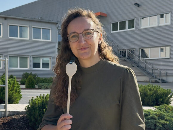 Frau mit lockigen Haaren und Brille hält einen Holzlöffel in der Hand vor einem modernen Gebäude.